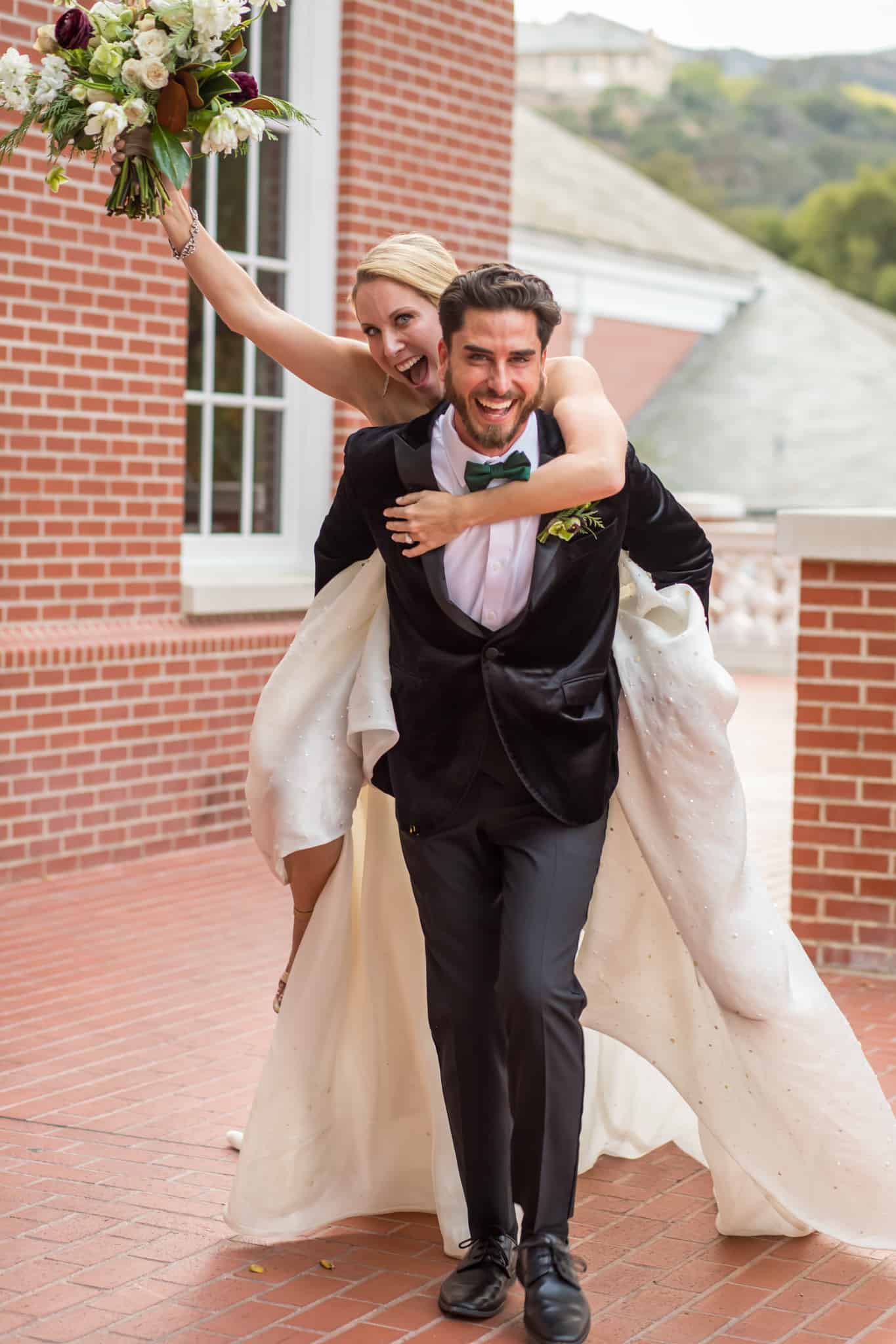 candid wedding photo off bride on grooms back as he carries her over a patio