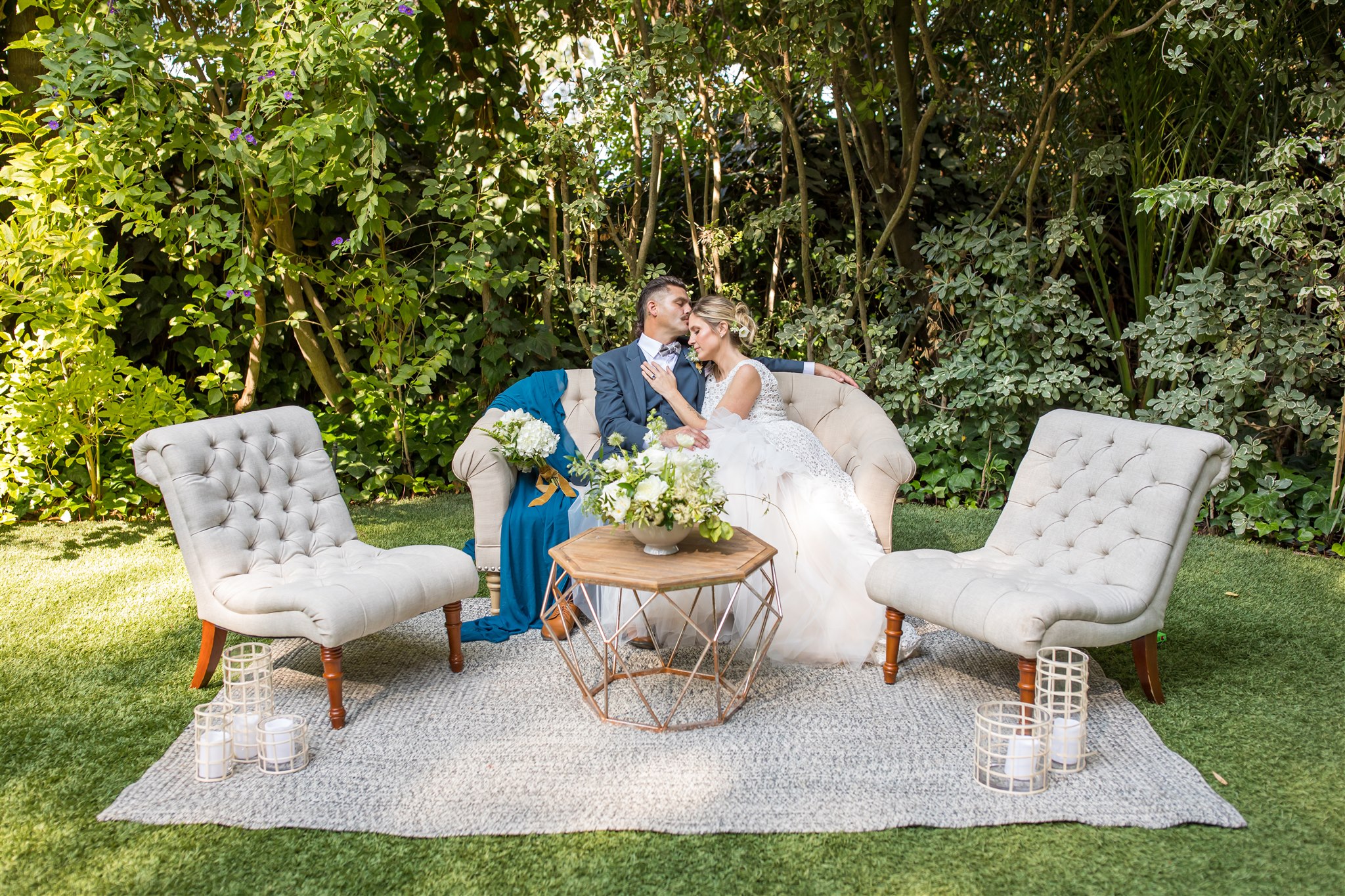 outdoor wedding lounge set with bride and groom cuddling on a couch as the groom kisses the brides forehead