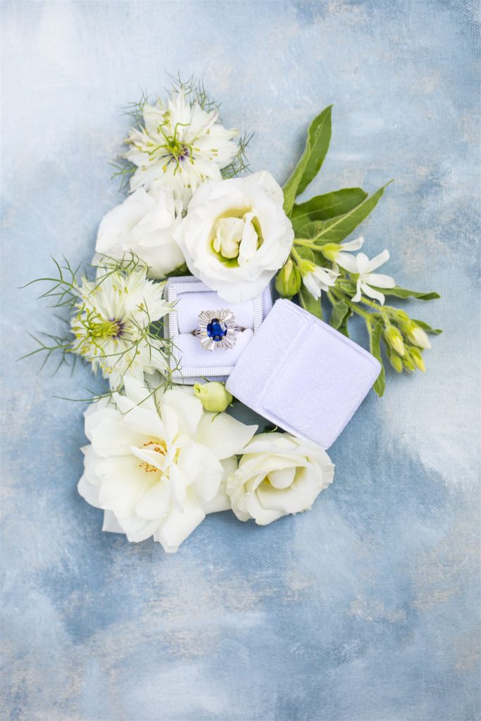 blue sapphire engagement ring in a white box with white wedding florals flat lay