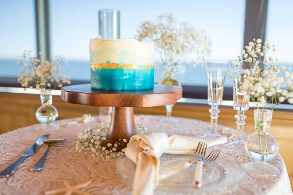 beach wedding cake that has blue and gold frosting sitting on a wood cake stand with florals surrounding