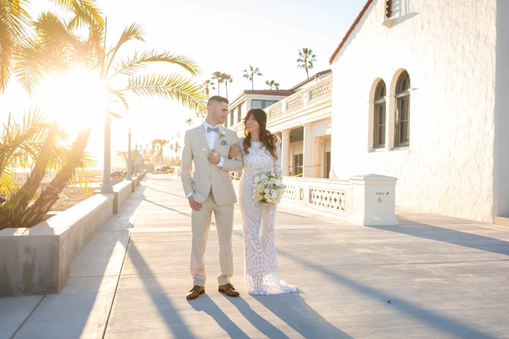 bride holding her grooms arm while looking up at him and smiling as the sun shines behind them through a palm tree