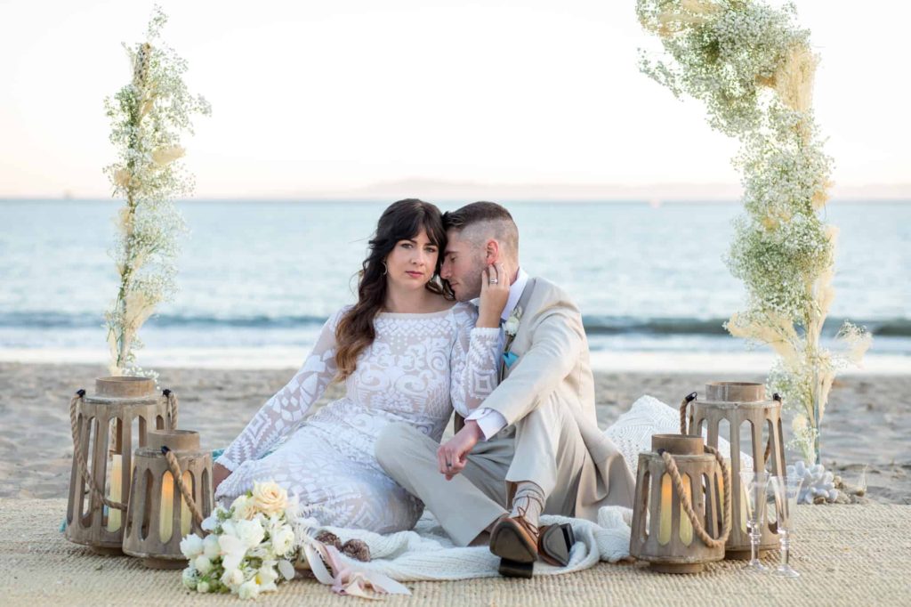 bride and groom sitting on a jute rug on the beach at Santa Barbara surrounded by wood lanterns and the ocean behind them