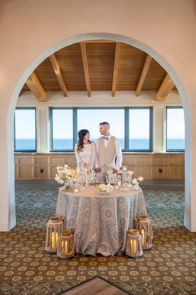 bride and groom standing next to their table at the wedding reception as they look romantically at one another while standing under an arch