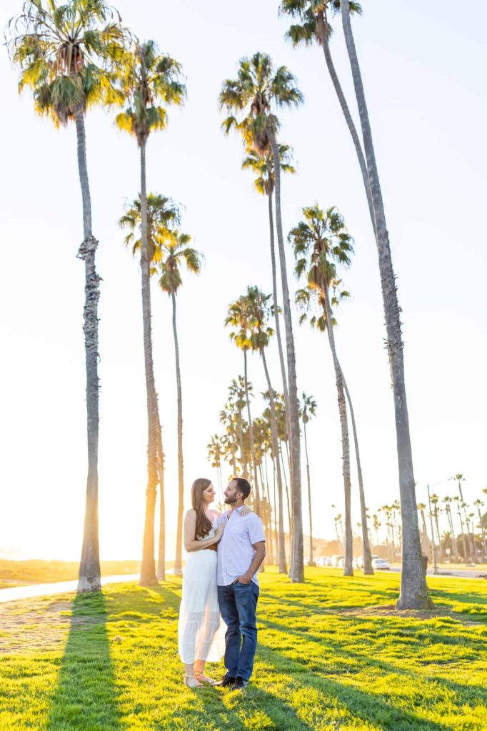 sunsetting over a green lawn with palm trees as an engaged couple embraces and smiles at one another