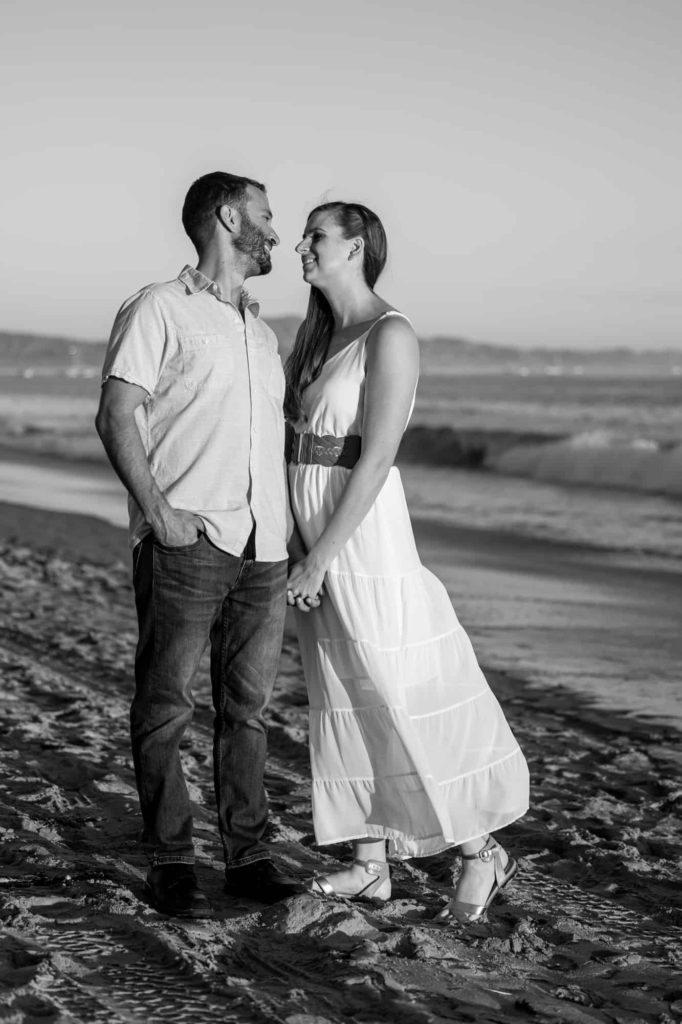 black and white engagement picture with woman holding her fiences hand and leans into him and smiles while the waves from the ocean crash over the beach behind them