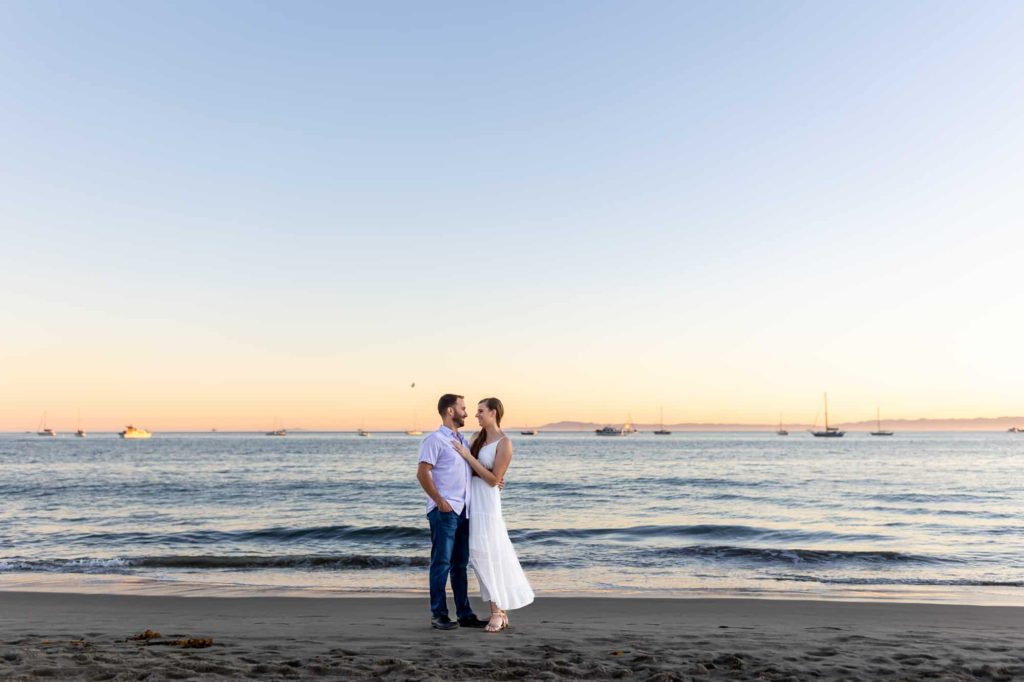 beach engagement photos with couple embracing on the sand while sail boats sail behind the couple as the sun sets
