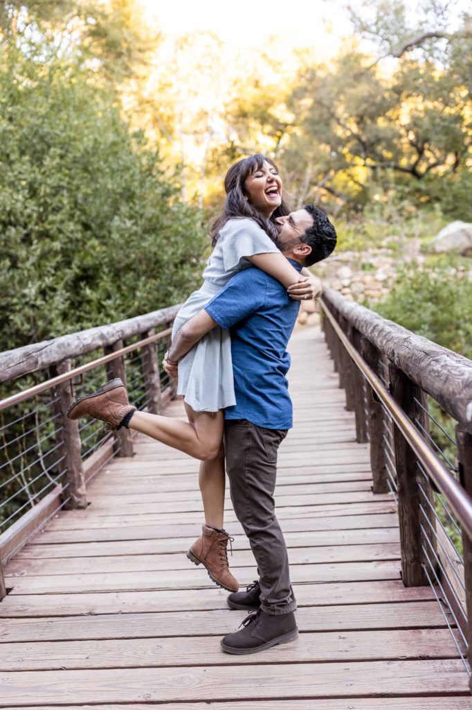 engagement photos on a bridge in Santa Barbara Botanical Gardens as the man picks up his fiance and they laugh together