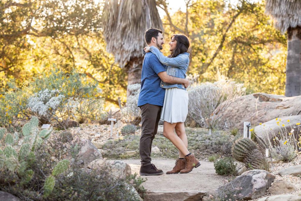 summer desert engagement photos in Santa Barbara with man and woman embracing each other as they smile with cacti and rocks surrounding them
