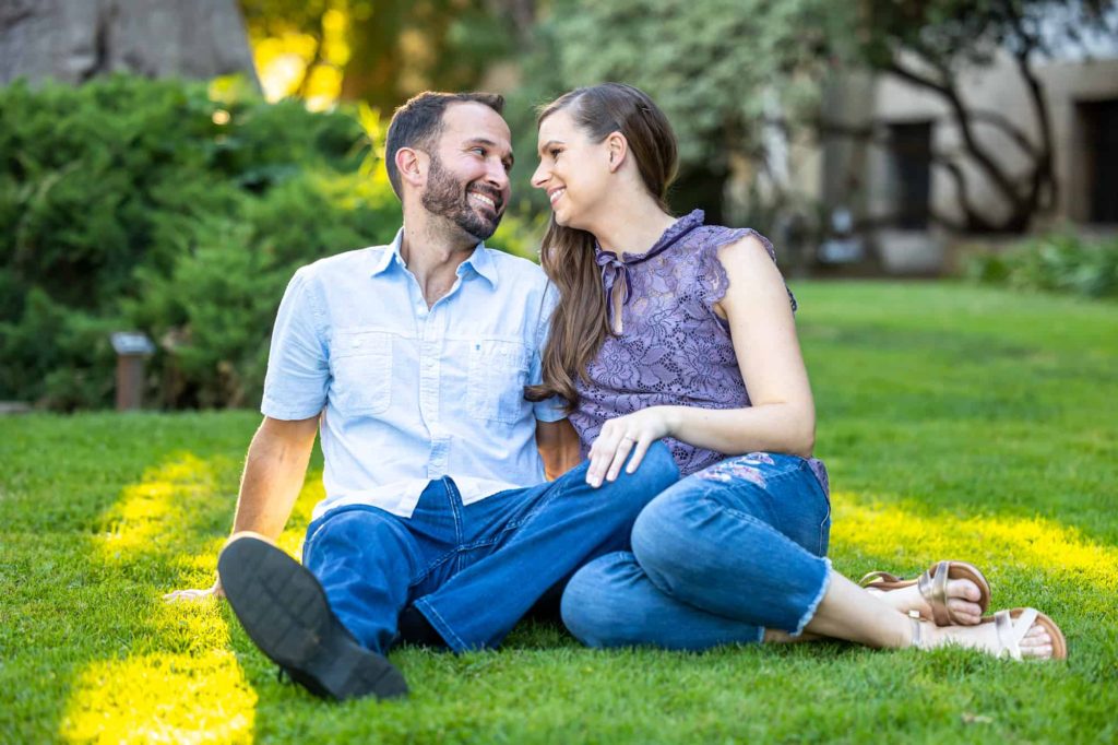 couple lounging on the lawn as the sun shines over them as they look at one another and smile during their engagement photoshoot