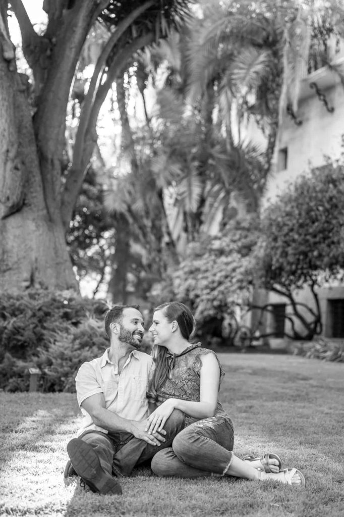black and white engagement photo with man and woman sitting on a lawn in the gardens as they hold hands and smile at each other