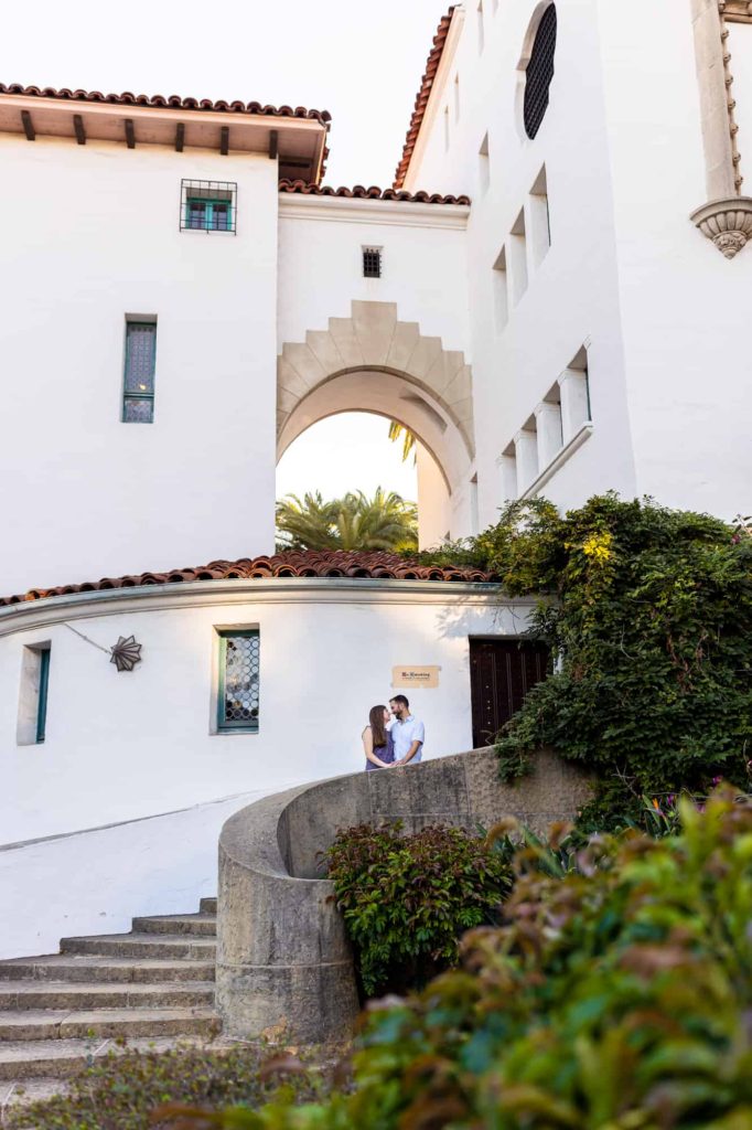 engaged couple standing on the stairs of the Santa Barbara courthouse while holding hands and looking at each other
