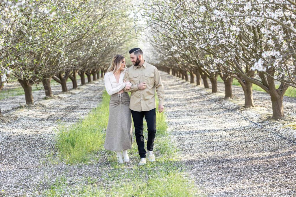 spring engagement pictures with man and woman walking as the woman holds the mans arm with almond blossoms surrounding them