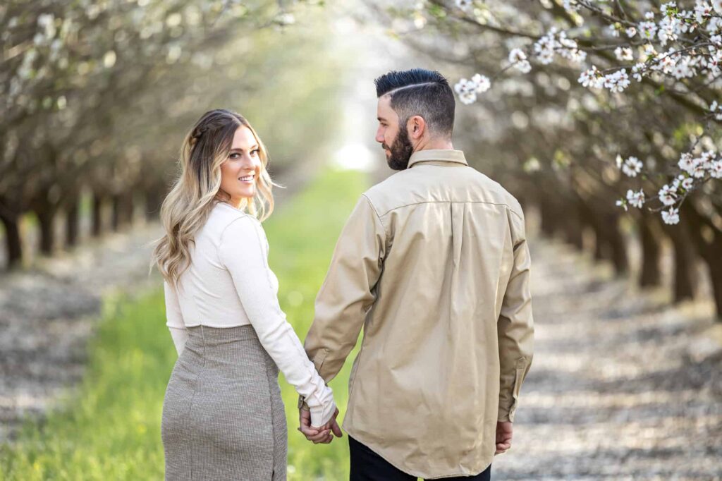 engaged couple holding hands and walking down a green grass aisle thats lined with almond tree blossoms