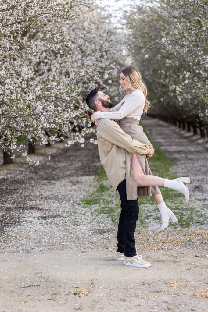 man picking up his fiance as she kicks up her foot and smiles down at the man in an almond orchard