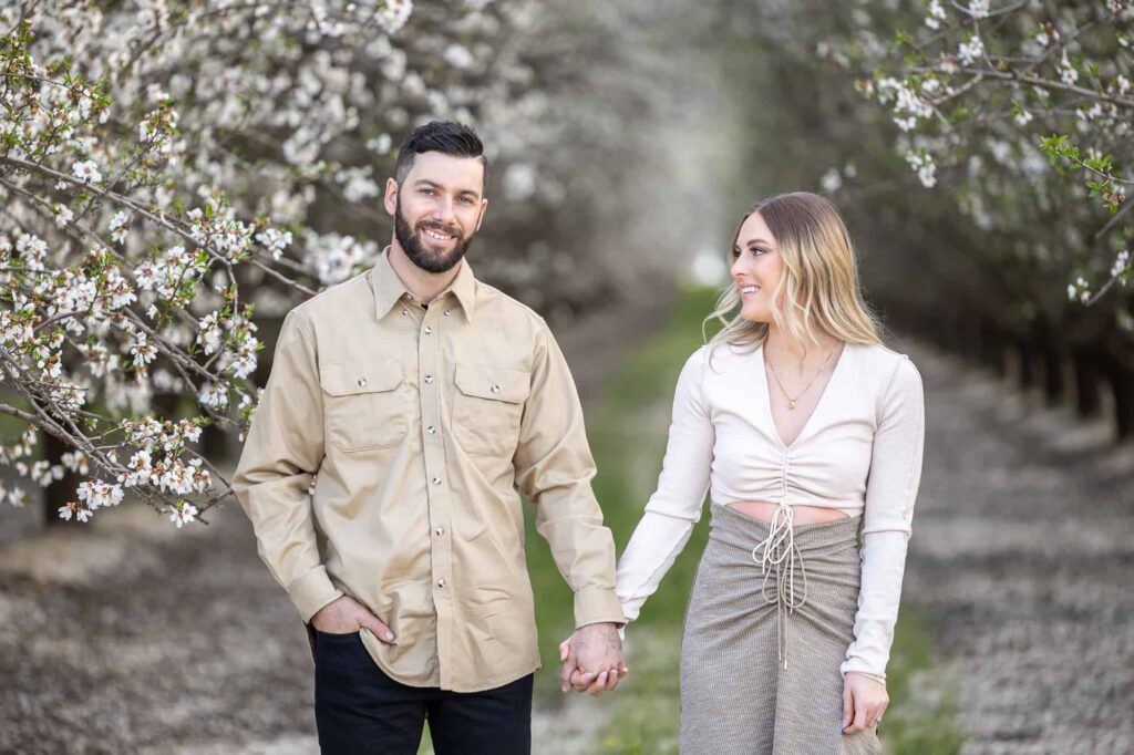 spring engagement pictures with man and woman holding hands and smiling while walking through a blossoming orchard