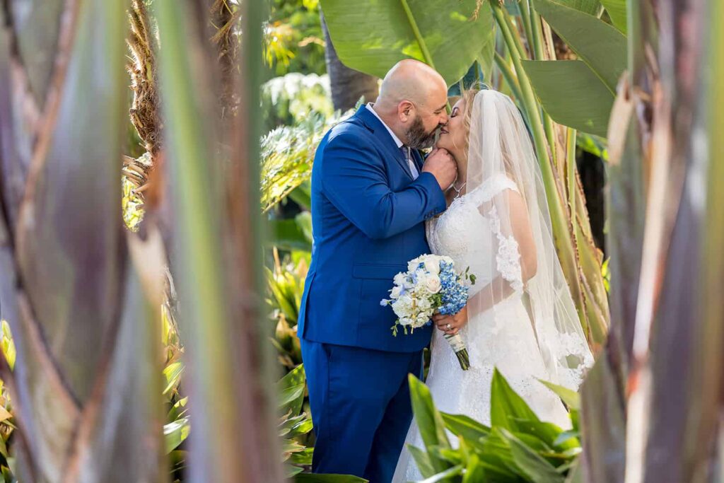 wedding photo in a garden full of tropical trees and plans with bride and groom leaning into one another as they kiss