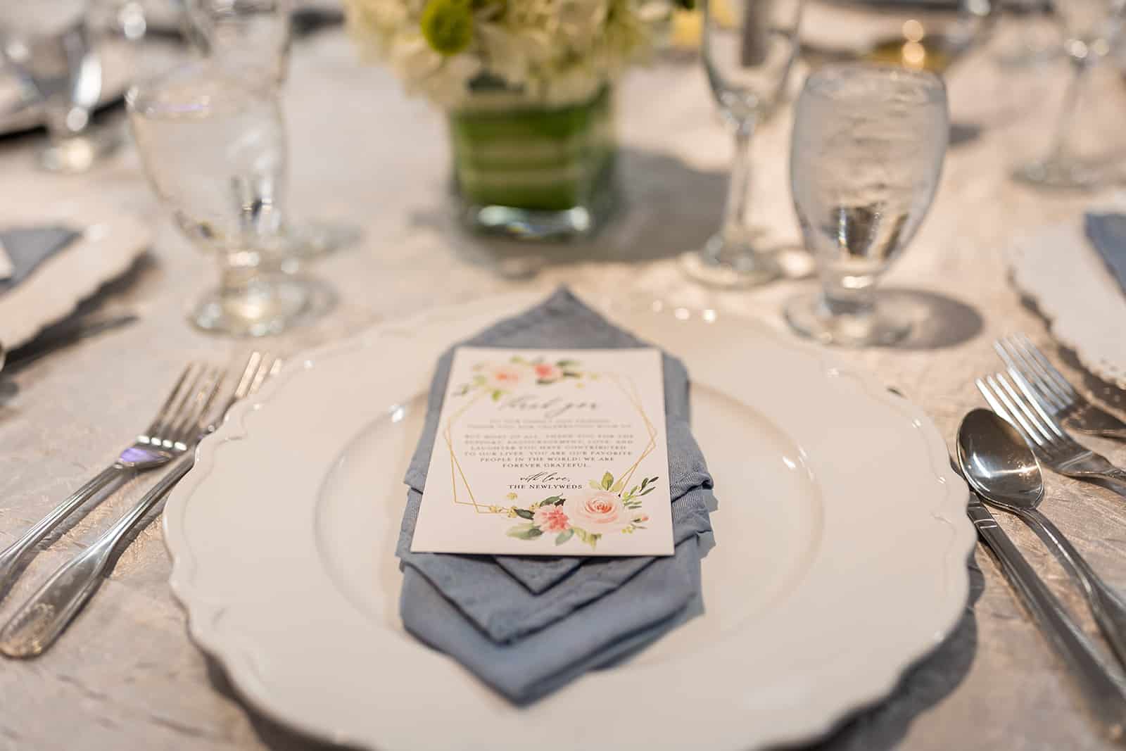 wedding reception dinner setting with a white plate, silver flatware, dusty blue napkins and a thank you card