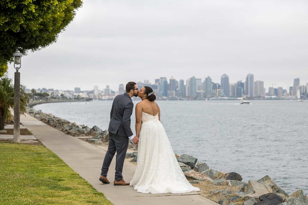 bride and groom kissing on a path by the beach with san diego skyline in the background