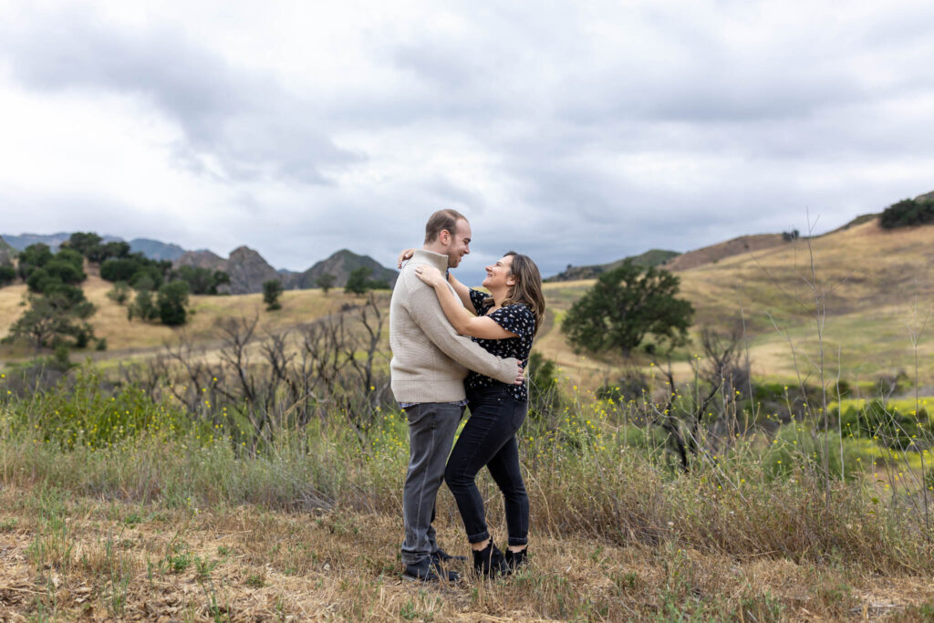 man holding woman around the waist as she holds him around the shoulder in a field on a cloudy day for their Malibu Creek State Park Engagement Photos