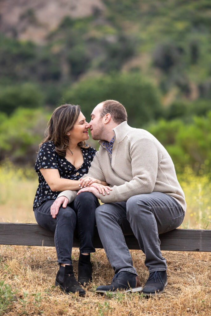 outdoor California engagement photos with man and woman sitting next to each other on a low fence in a field in Malibu State Park as the man leans in to kiss his fiance for their summer engagement session