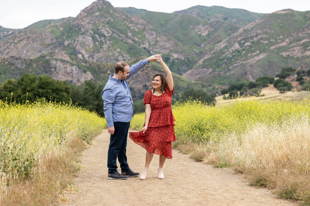 man and woman dancing for their outdoor California engagement photos on a dirt path as the man twirls the woman who is in a red dress withe the Santa Monica mountains in the distance