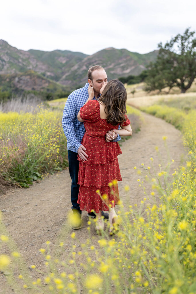 man kissing a woman on a dirt path in California State Park with yellow flowers in the field with man in a blue shirt and woman in a red dress