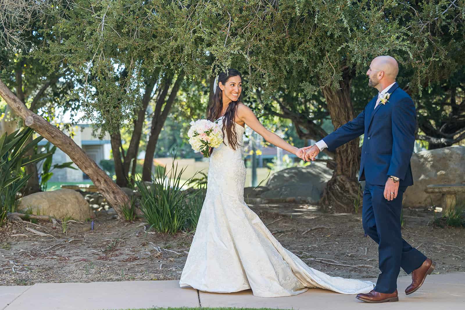 bride holds grooms hands and leads him through a garden at their Ventura wedding venue