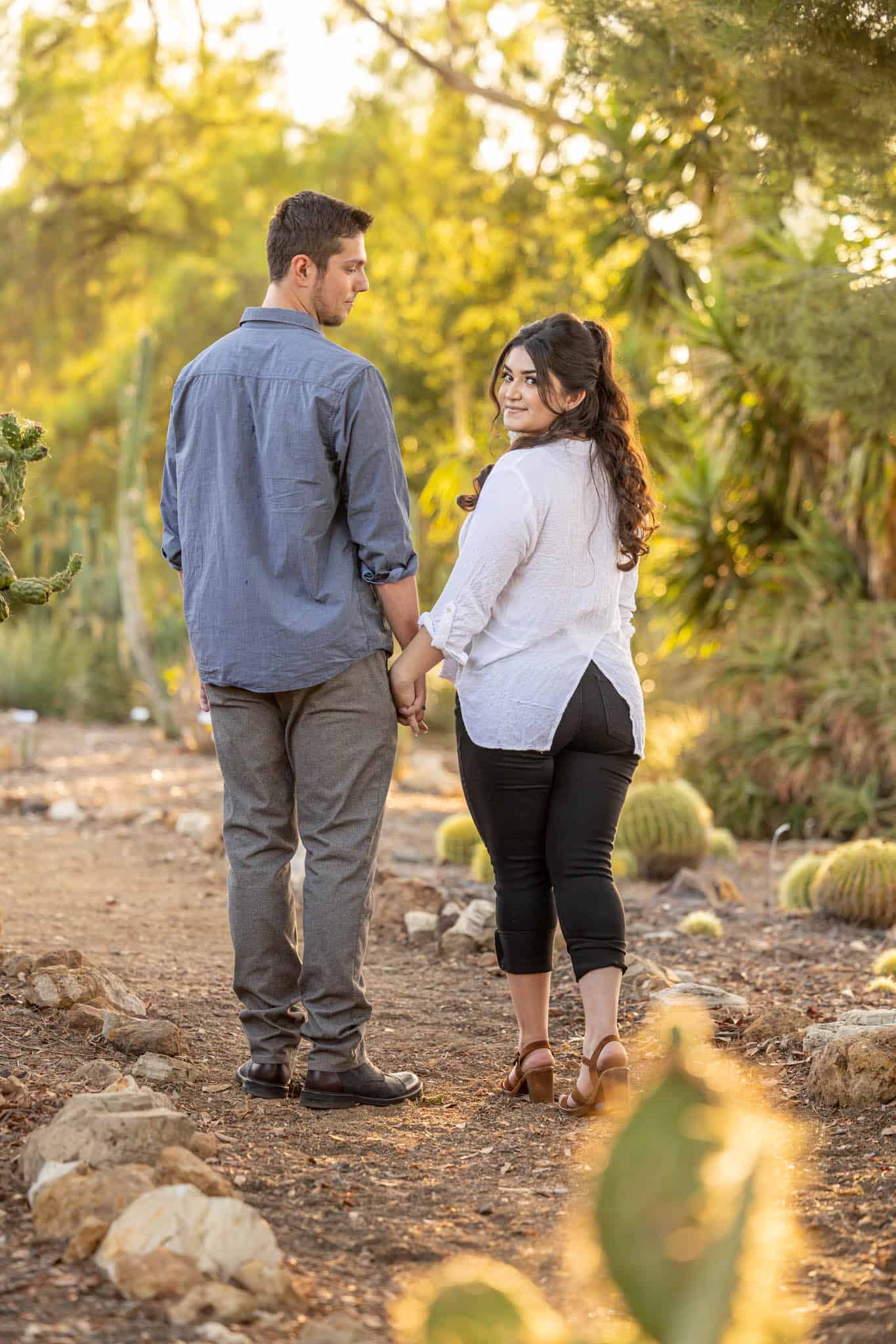 Thousand Oaks wedding photographer captures engagement session in Conejo Botanical Gardens with man and woman walking down a path together as the woman looks over her shoulder