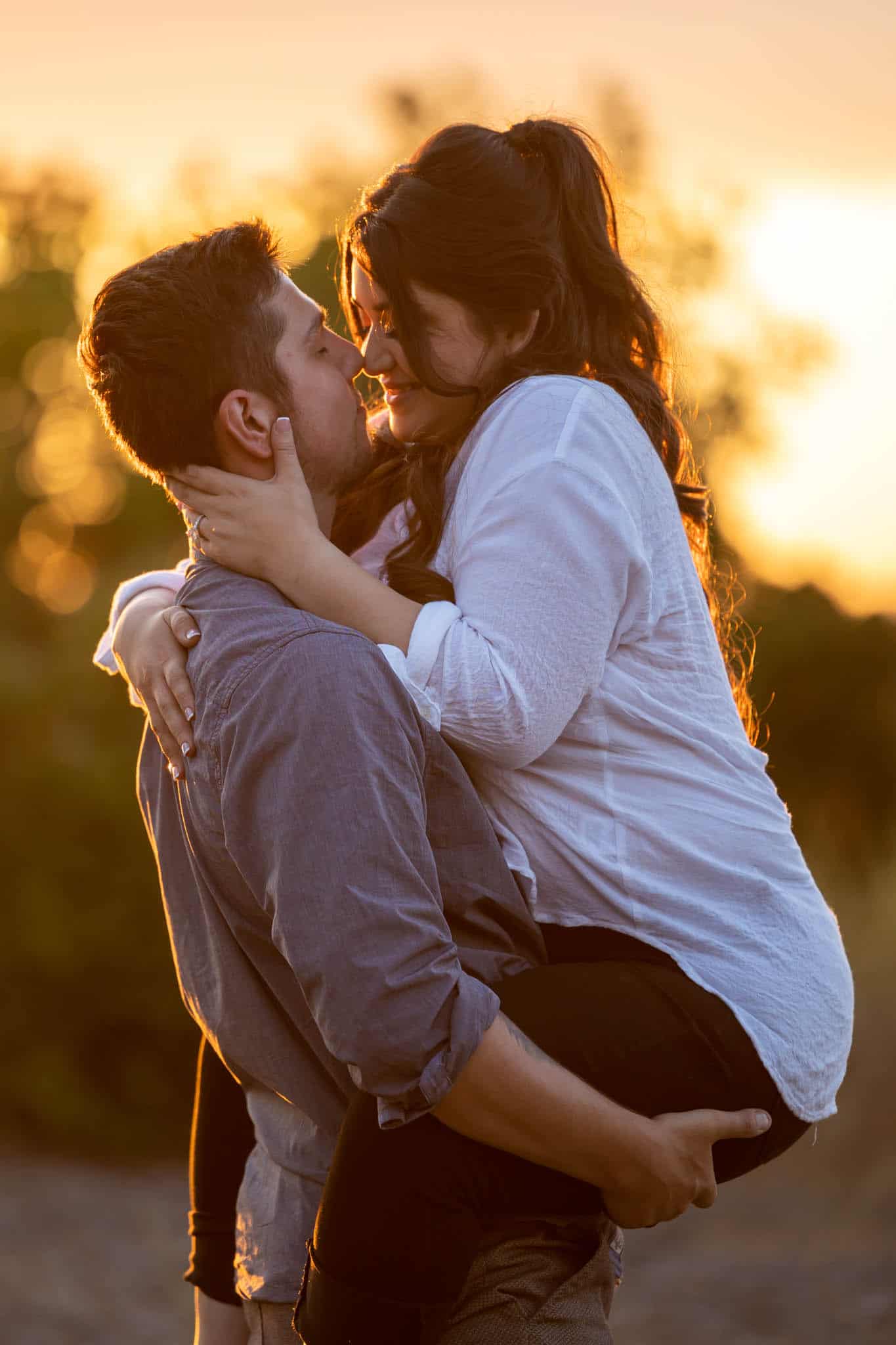 woman jumps up as her fiance holds her as she holds his face and they lean into kiss each other