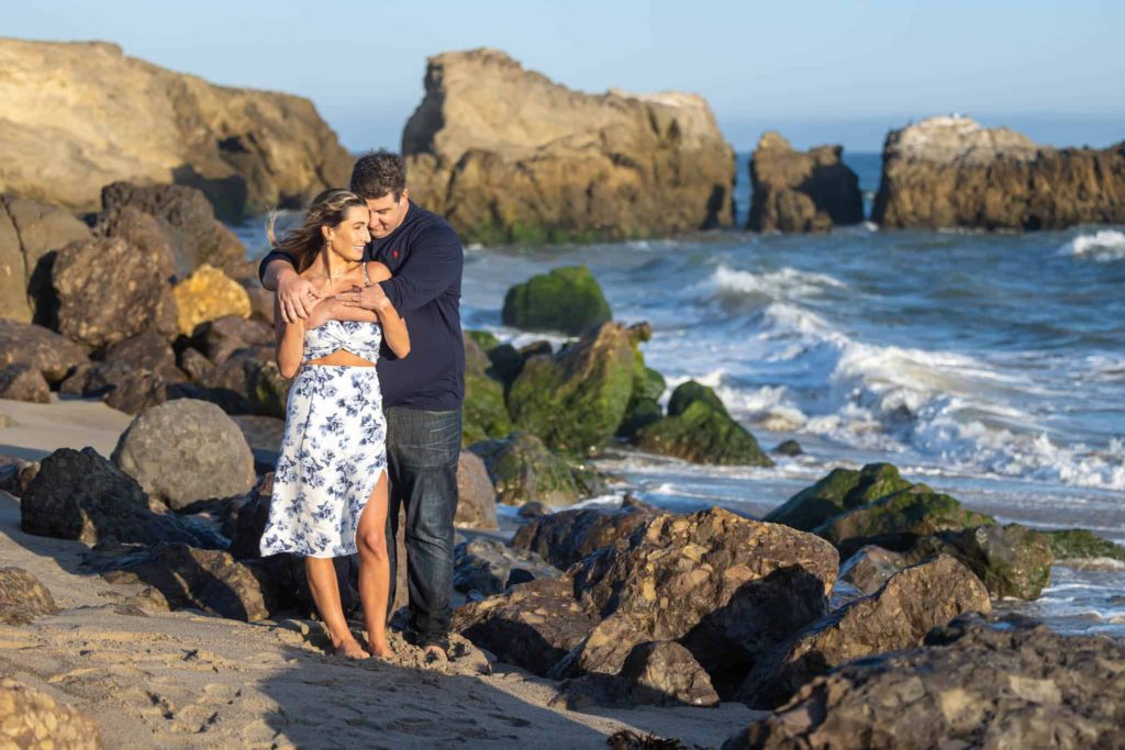 man and woman embracing on the rocks at the beach for engagement pictures by Los Angeles wedding photographer