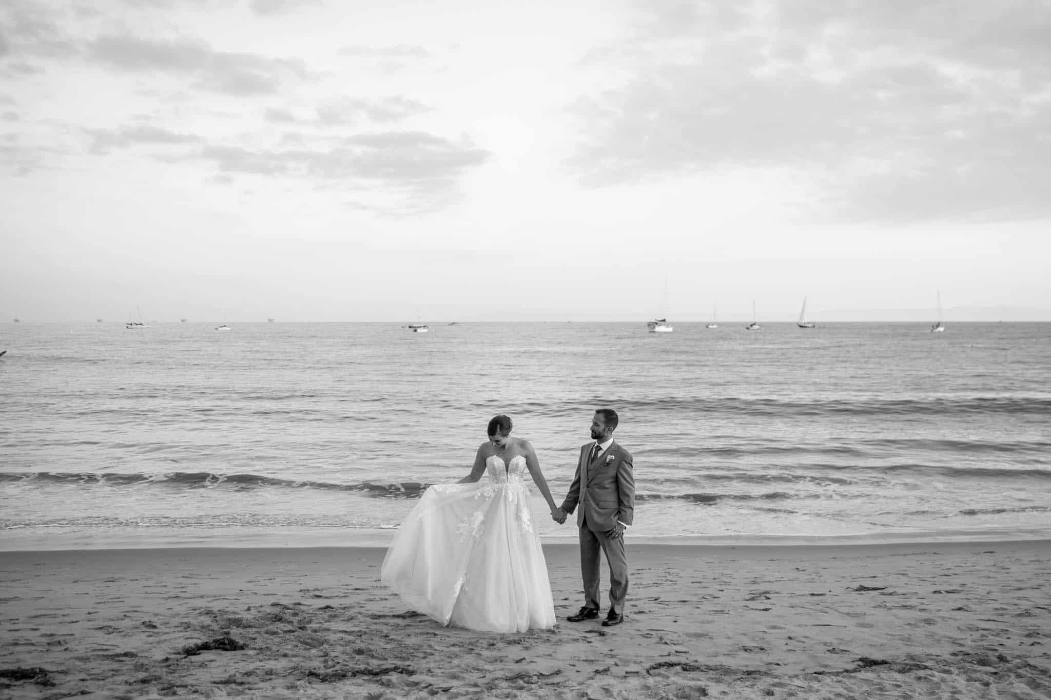 Santa Barbara wedding photographers capture bride and groom walking on the beach together while holding hands with boats int he distance while the bride plays with her dress