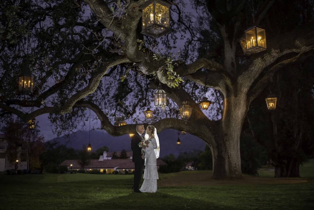 night time dance with bride and groom under a large tree with lantern hung as the sun sets over the mountains in the distance at Ojai Valley Inn with Ventura county wedding photographer